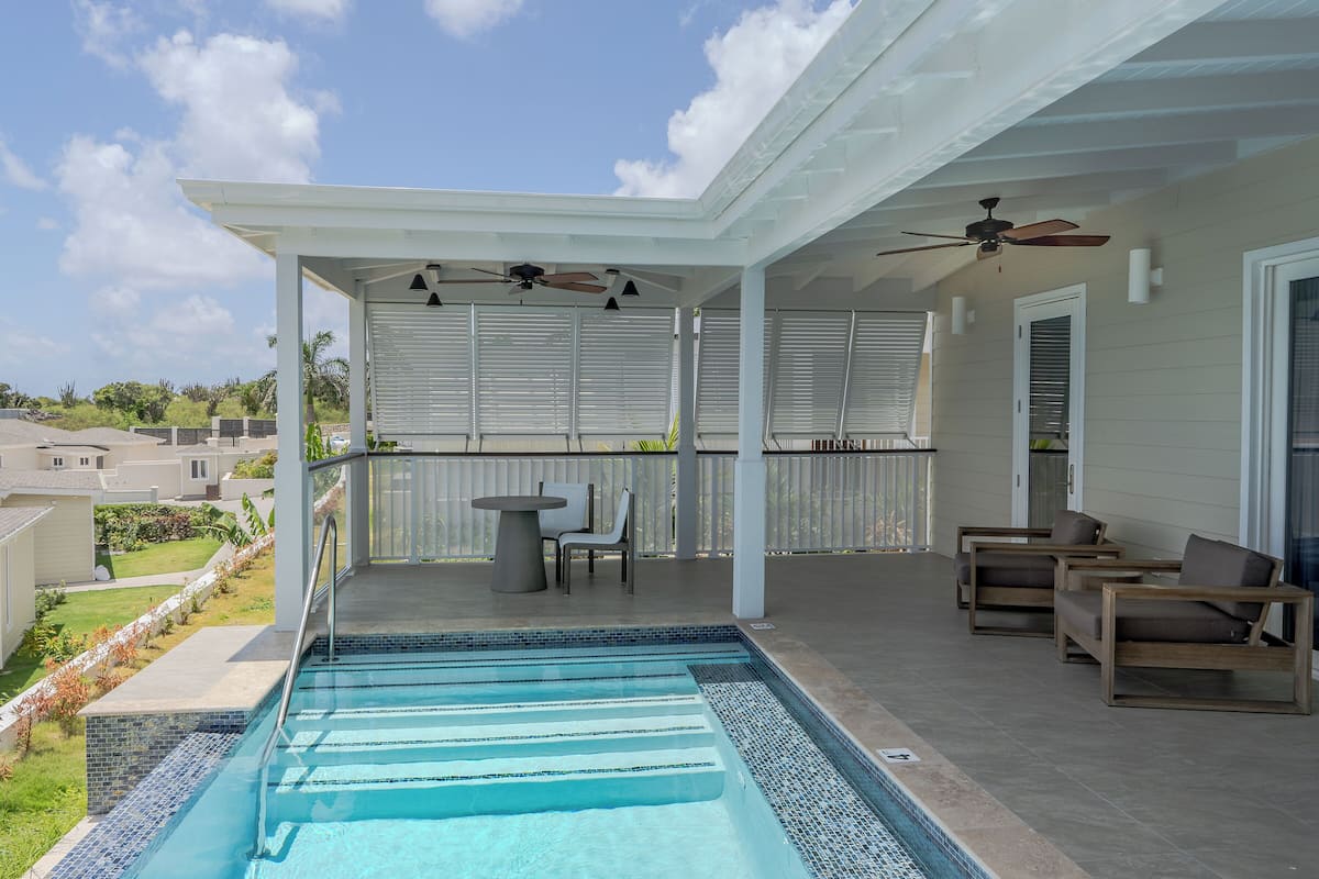 A covered outdoor swimming pool area with a patio, seating, and a view of the neighborhood.
