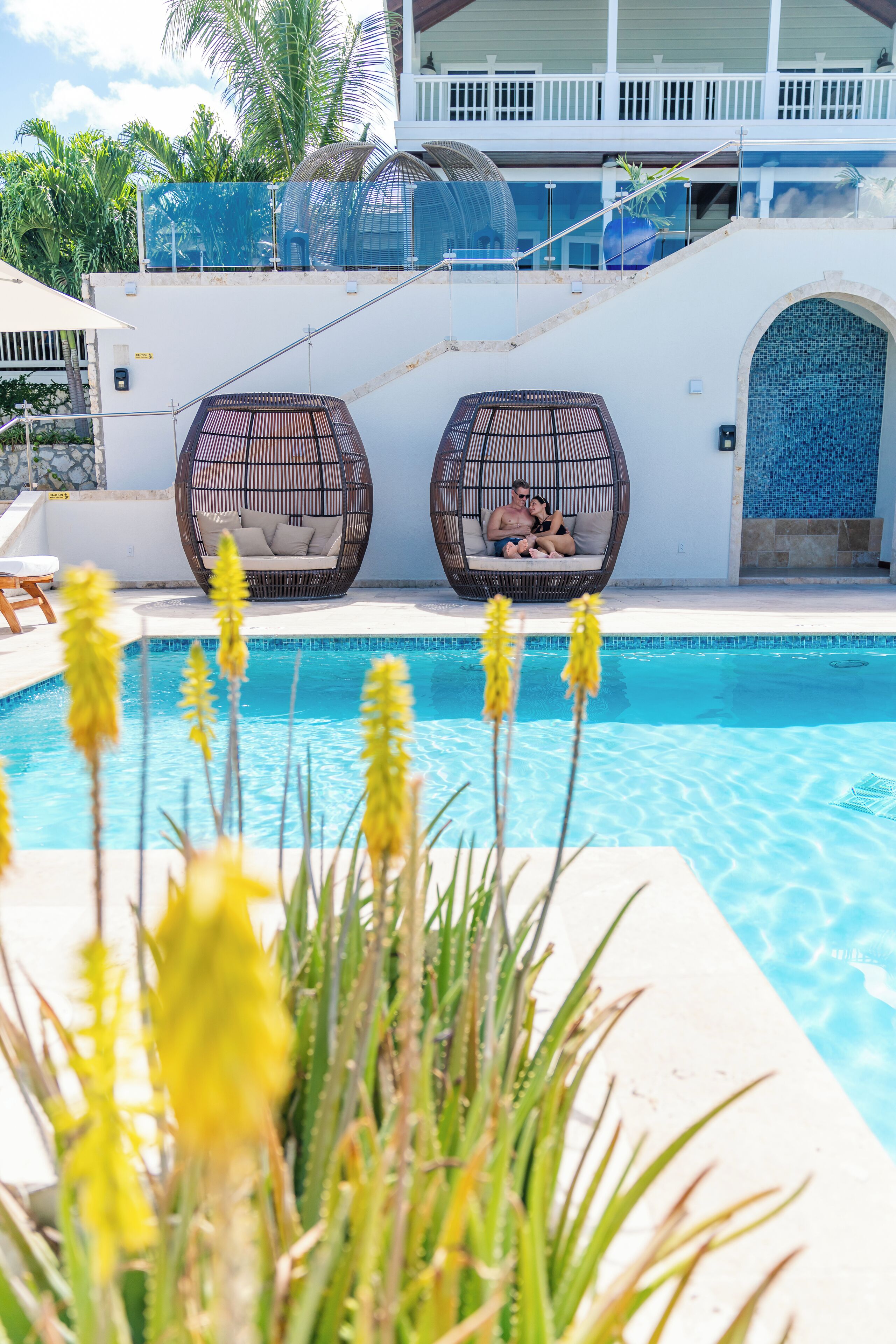 A pool area with two wicker chairs, a white building with a balcony, and tropical plants.