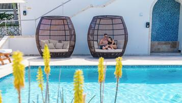 A pool area with two wicker chairs, a white building with a balcony, and tropical plants.