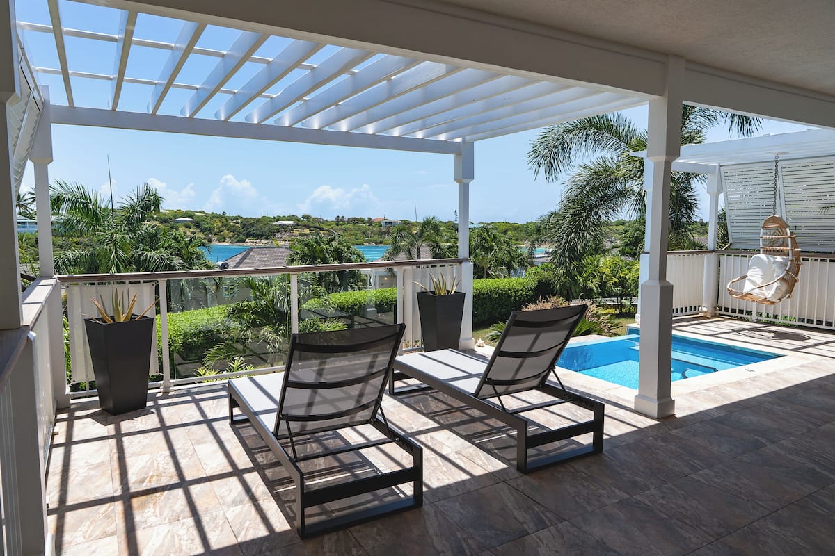 A covered outdoor area with a pool, two lounge chairs, and a view of a tropical landscape.