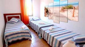 A room with three single beds, each with striped bedding and towels. There is a beach-themed wall art above the beds, and a window with orange curtains allowing natural light. - GBH Volcanic Beach (La Oliva)