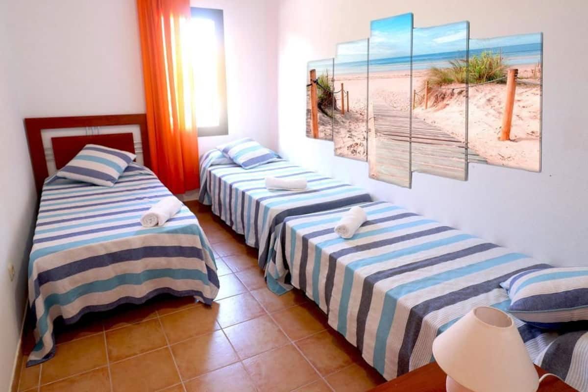 Quarto com três camas de solteiro, cada uma com roupa de cama e toalhas listradas. Há uma obra de arte com tema de praia acima das camas, e uma janela com cortinas laranja que permite a entrada de luz natural.