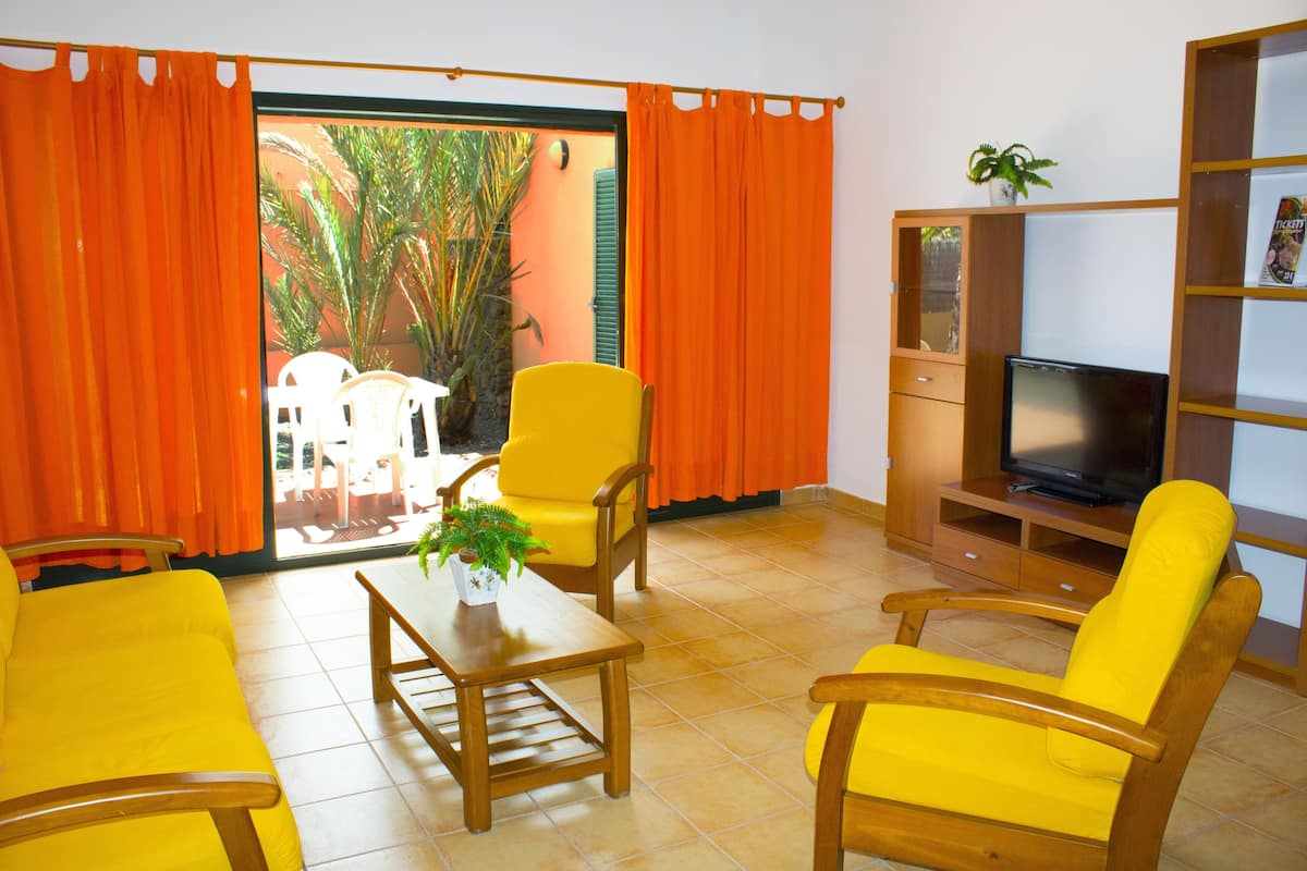 Sala de estar com móveis amarelos, uma televisão e vista para um jardim através de uma porta de correr.