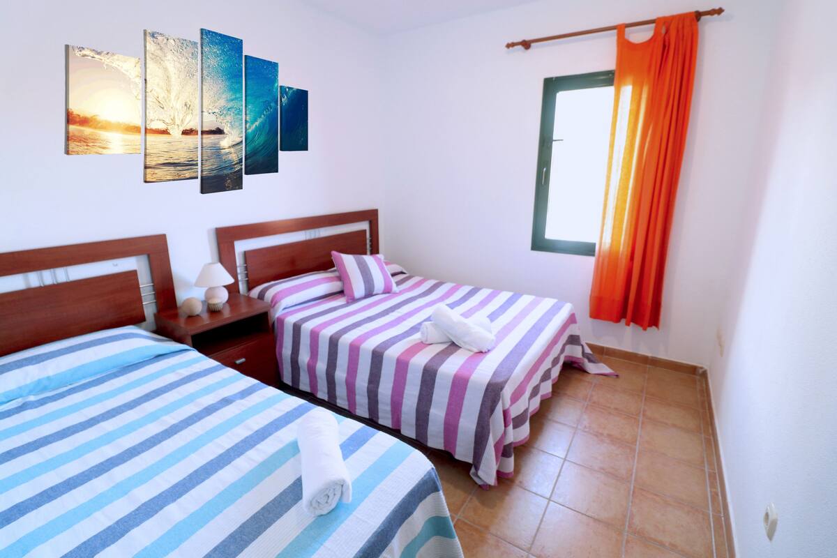 Quarto de hotel com duas camas, uma janela com cortinas laranja e uma parede com três quadros.