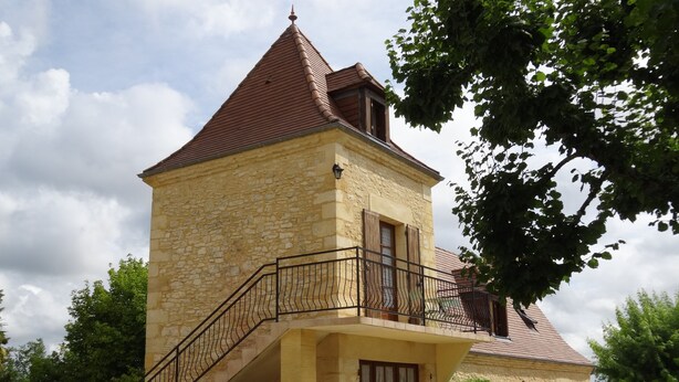 Ein Steinhaus mit rotem Ziegeldach, einem Balkon mit schwarzer Eisenbalustrade und einer Treppe zum Eingang.