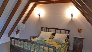 Ein Schlafzimmer mit schrägem Dach, einem Bett mit gemustertem Überwurf, zwei Wandleuchten, einem Nachttisch und einem kleinen Beistelltisch mit einer Vase.
