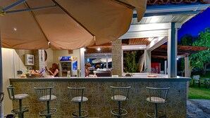 Un área de bar al aire libre con mostrador de baldosas de piedra, taburetes de metal y sombrillas que dan sombra.