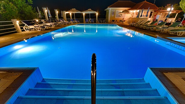 Una piscina con sillas de descanso y sombrillas, rodeada por una terraza de madera y un edificio con tejado de azulejos.