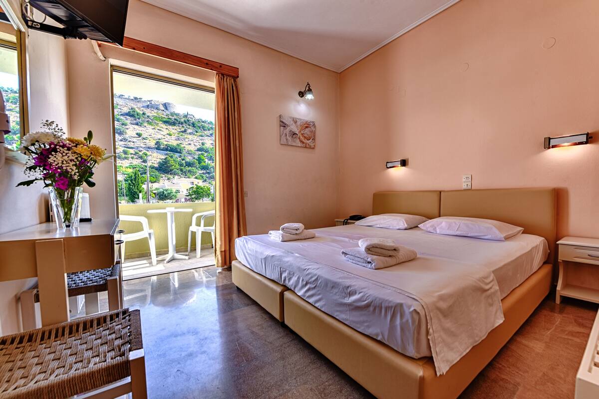 Une chambre d’hôtel avec un grand lit, un bureau avec une chaise, un vase de fleurs et une vue sur une colline.