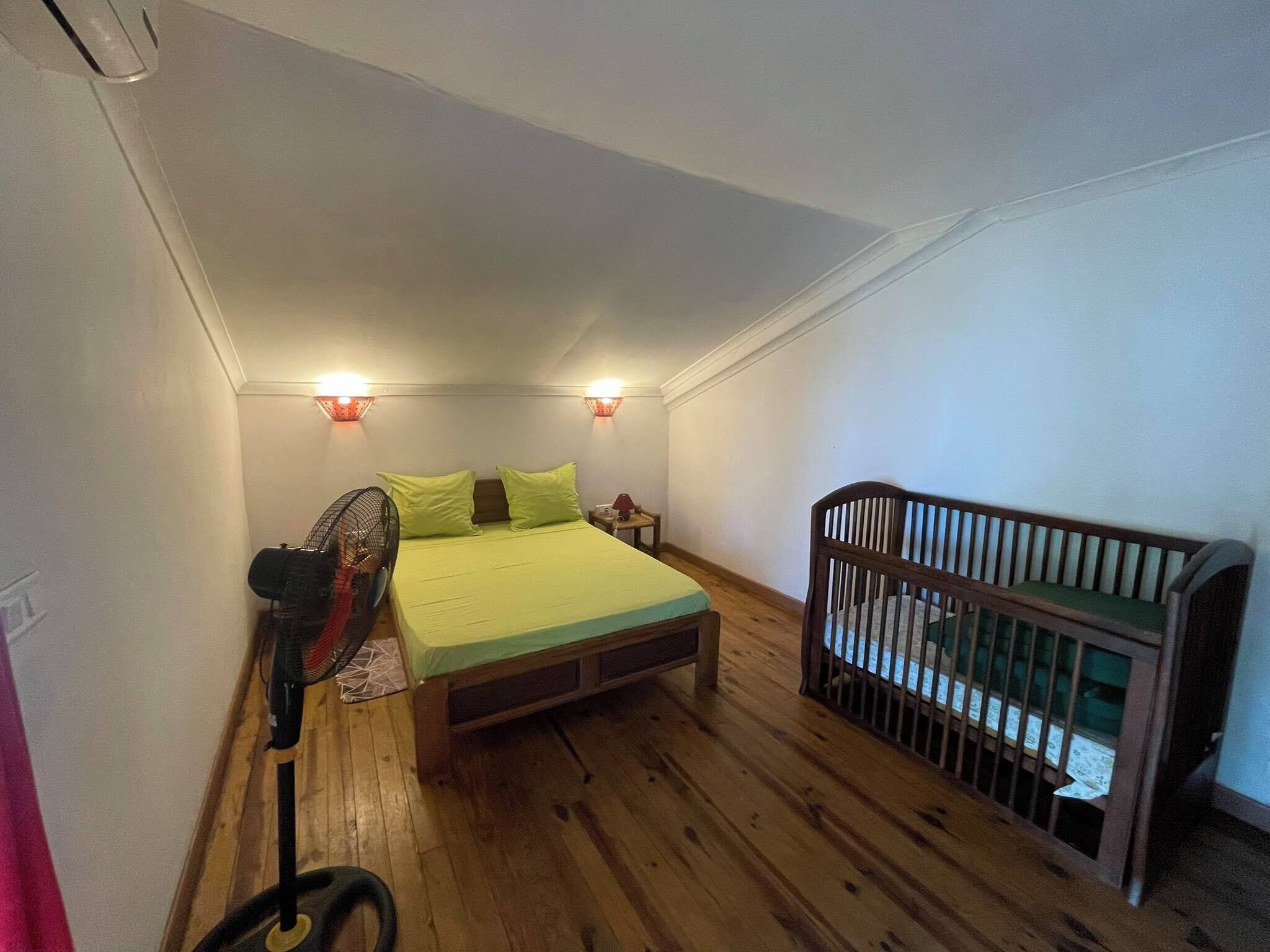 A small, single-room apartment with a wooden floor, a bed with green bedding, a wooden crib, and two wall-mounted lights.