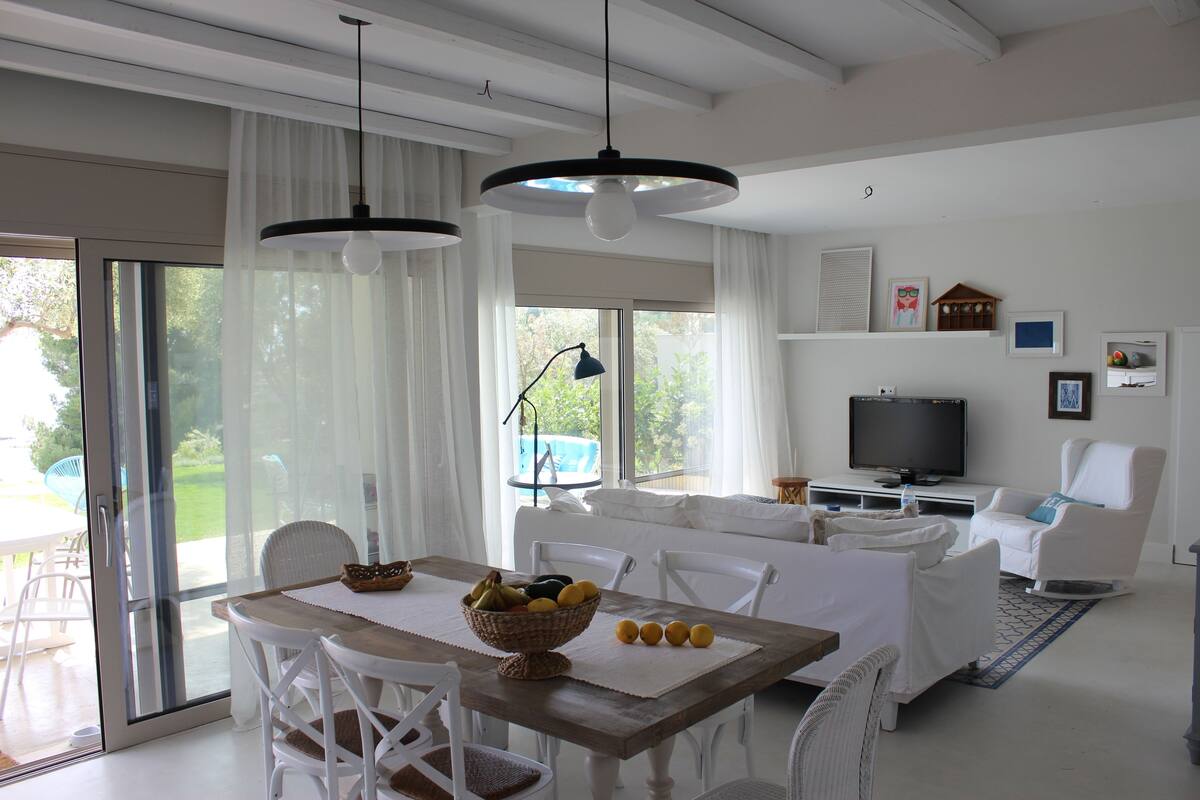 A modern living room with a dining area, white furniture, a television, and a large window with curtains.