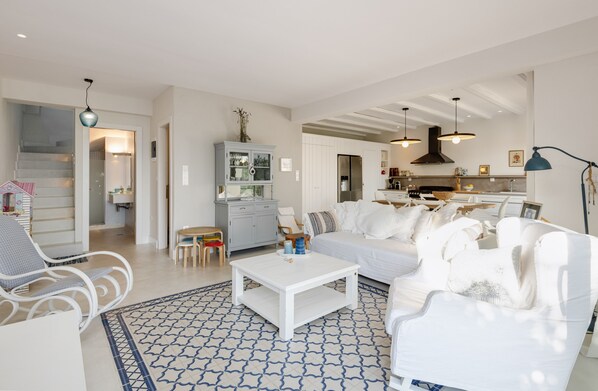 A spacious living room with a white sofa, a coffee table, and a patterned rug. A kitchen is visible in the background with modern appliances and pendant lighting. - Akti Elia Villas (Sithonia)