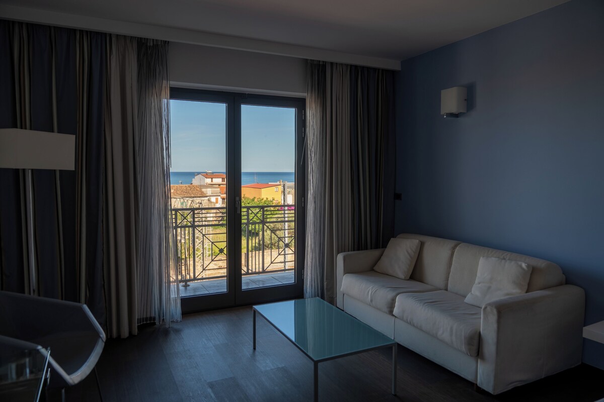 A room with a sofa, a glass coffee table, and a view of the sea through a sliding door.