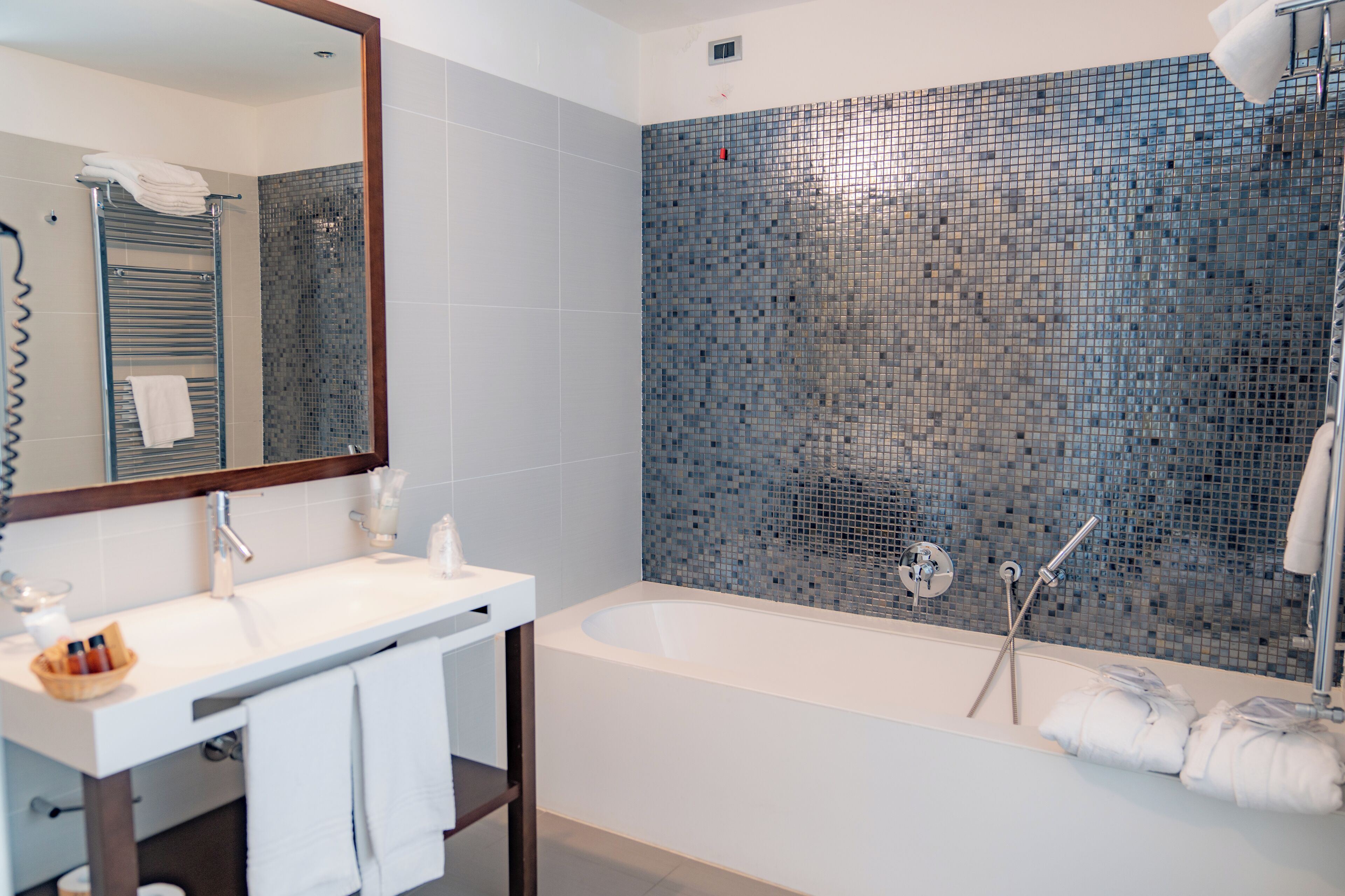Casa de banho moderna com lavatório branco, espelho e uma banheira grande com parede revestida em mosaico.