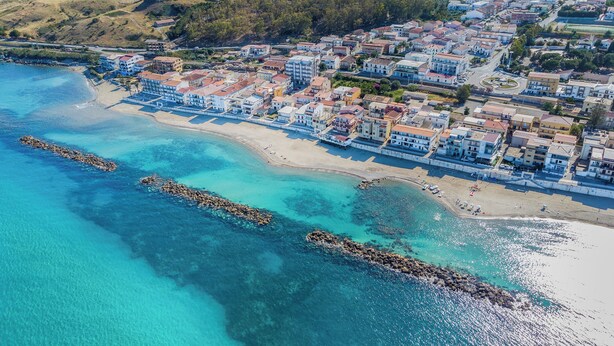 An aerial view of a coastal town with a beach, clear turquoise waters, and buildings along the shoreline. - Hotel Napoleon (Melissa)