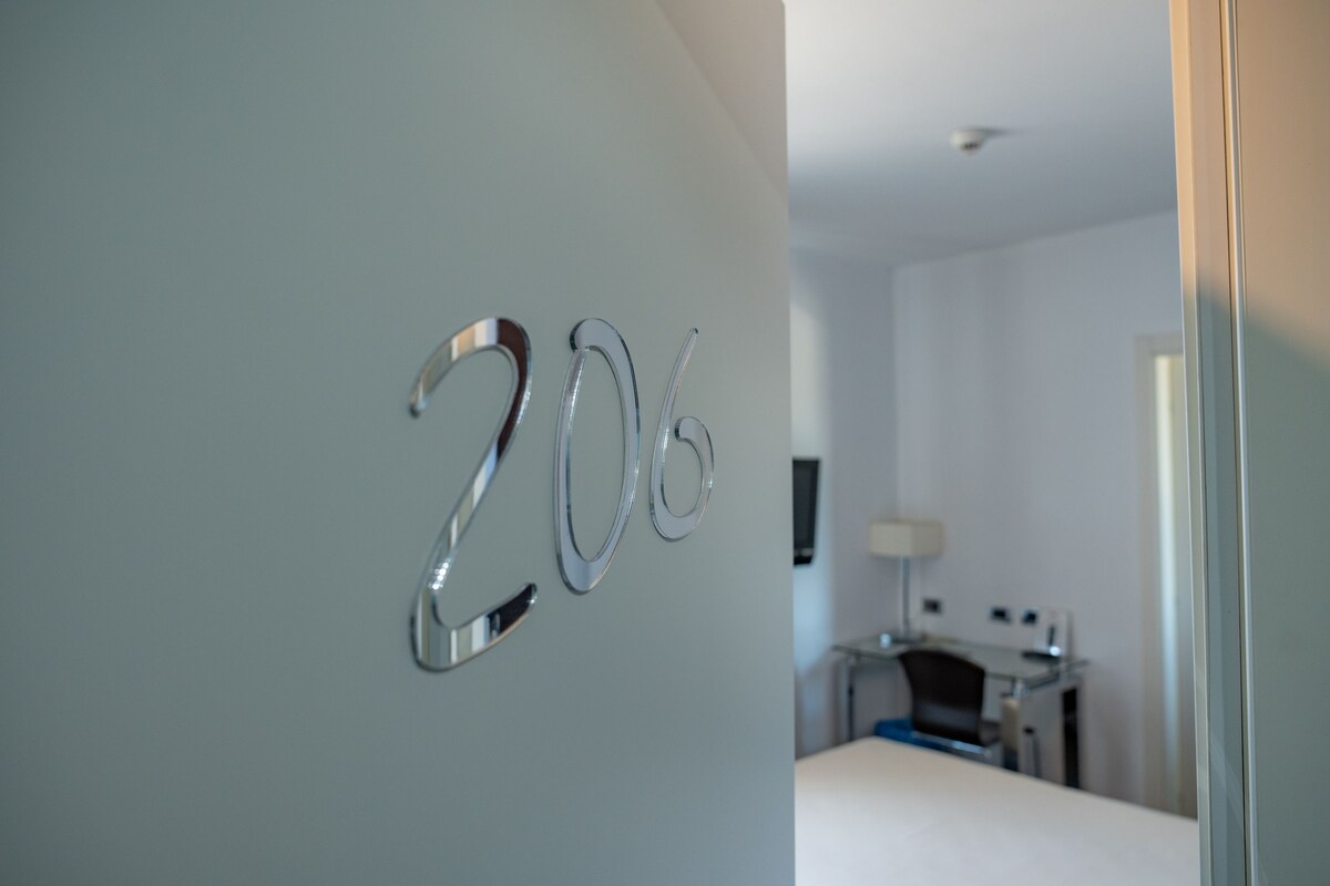 A hotel room with a number '206' on the wall, a bed, a desk, and a lamp.