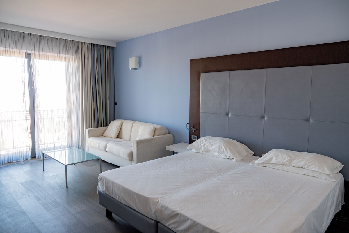 A hotel room with a bed, a sofa, a glass coffee table, and a view of the outside.