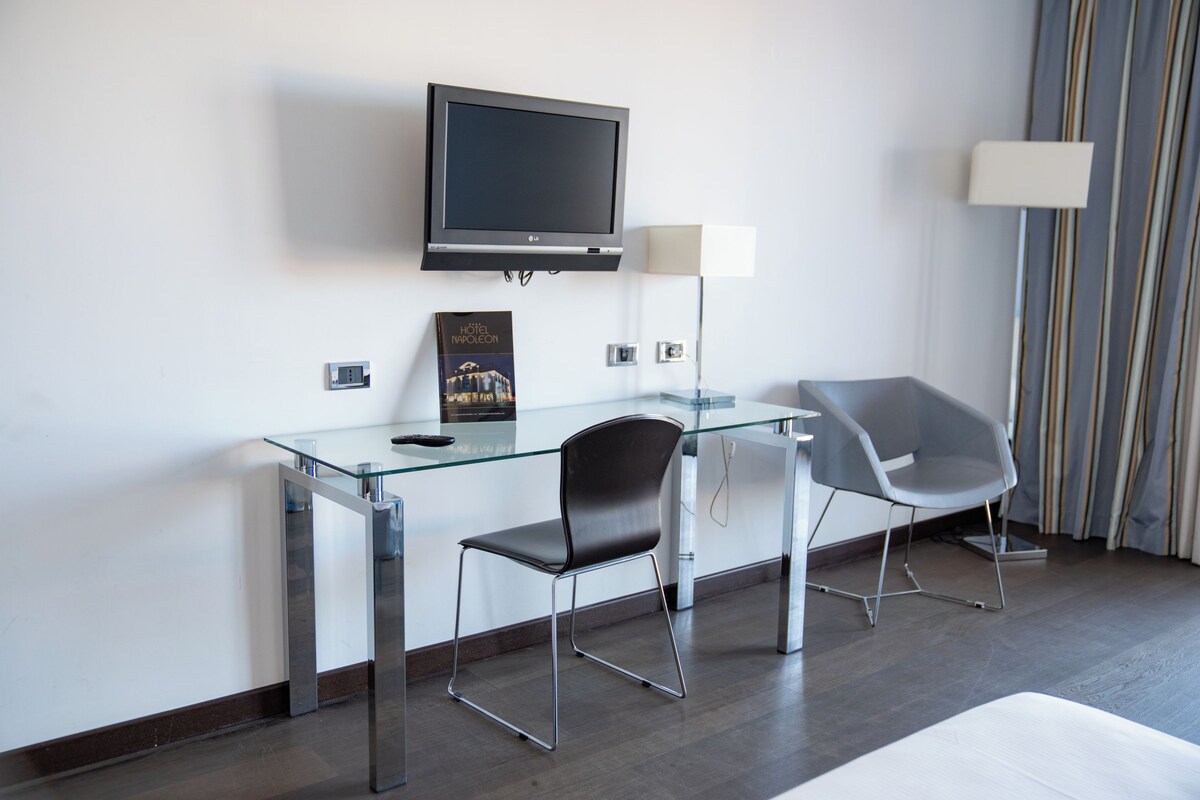 A modern hotel room with a glass desk, two chairs, a flat-screen TV, a lamp, and a poster on the wall.