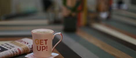 Una taza rosa con la leyenda “GET UP!” sobre una mesa de madera con periódicos y un posavasos.