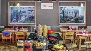 A buffet setup with various food items and beverages, tables with chairs, and two large windows with city views. - Ginger Surat City Centre (Surat)