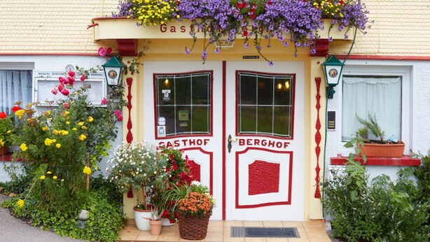 Un bâtiment doté de deux portes portant les inscriptions « Gasthof » et « Berghof », entouré de plantes et de fleurs en pot.