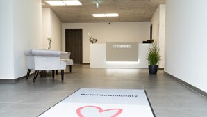 A hotel reception area with a reception desk labeled "Schlafplatz," a reception chair, a potted plant, and a heart-shaped rug on the floor. - Hotel Schlafplatz (Rodgau)