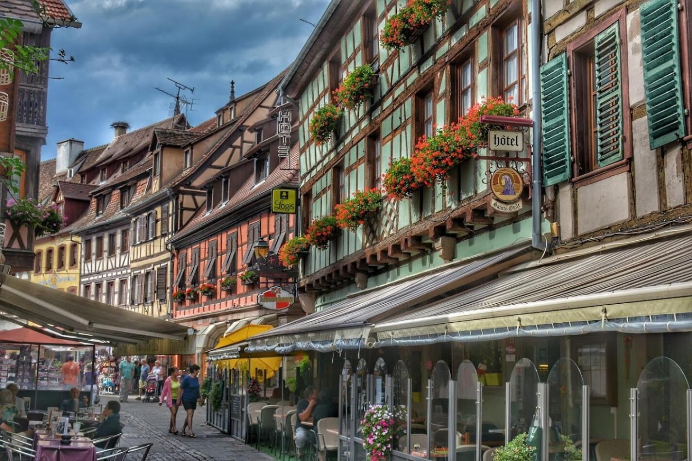 Una calle europea tradicional con un hotel que presenta un cartel, balcones con jardineras y una zona de estar al aire libre techada.
