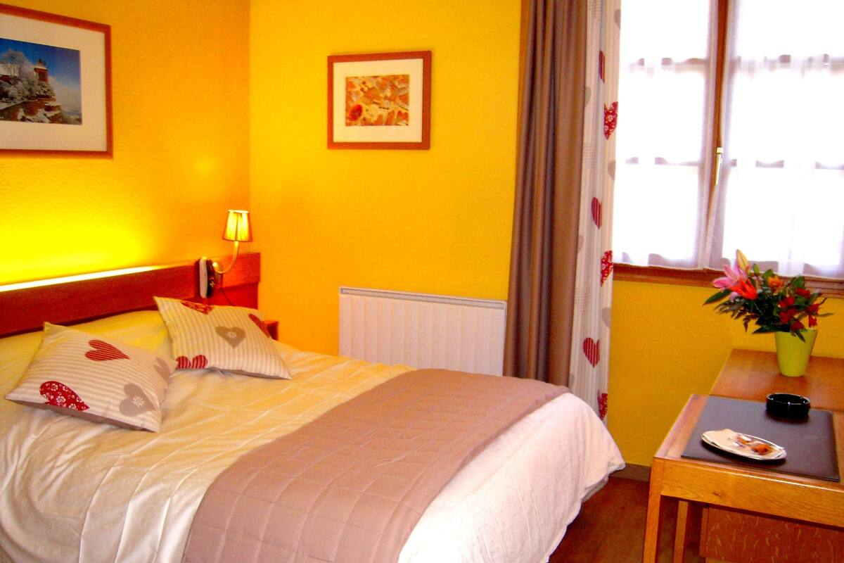 A hotel room with a bed, a nightstand with a lamp, a window with curtains, a small table with a vase of flowers, and a framed picture on the wall.