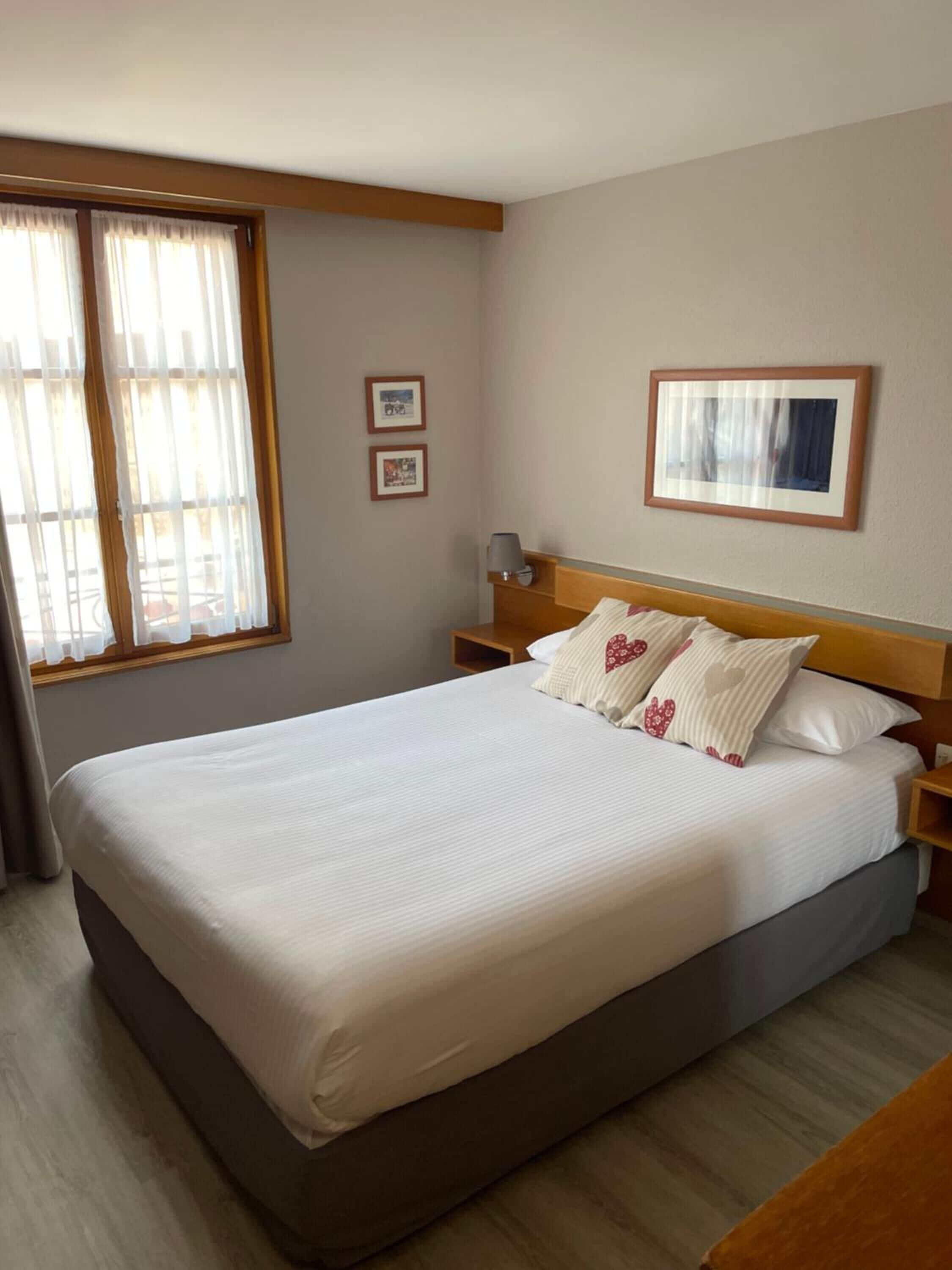 Una cama bien hecha en una habitación de hotel, con un cabecero de madera, una mesita de noche con una lámpara y dos cuadros en la pared.