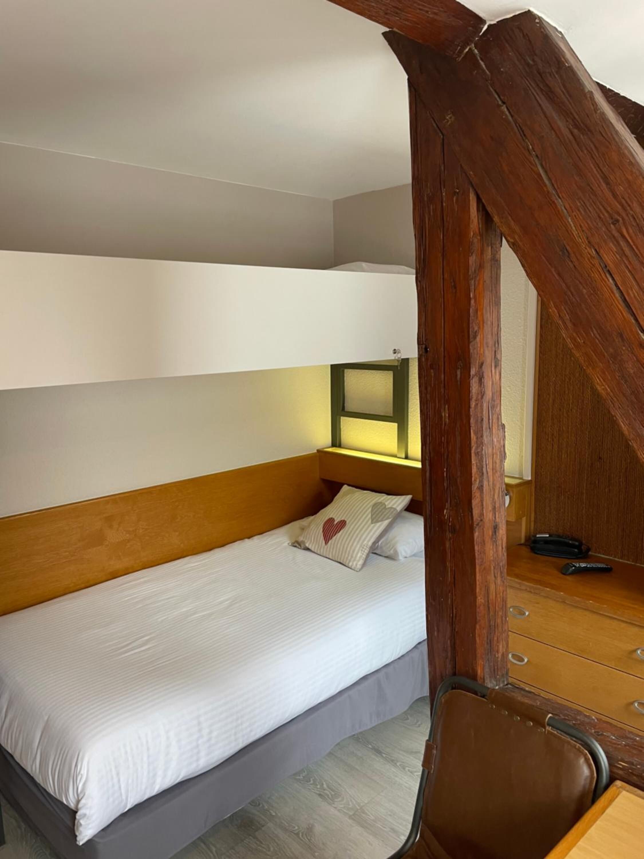 A single bed with a wooden headboard, a bedside table with a lamp, and a chair in a room with a sloped ceiling.
