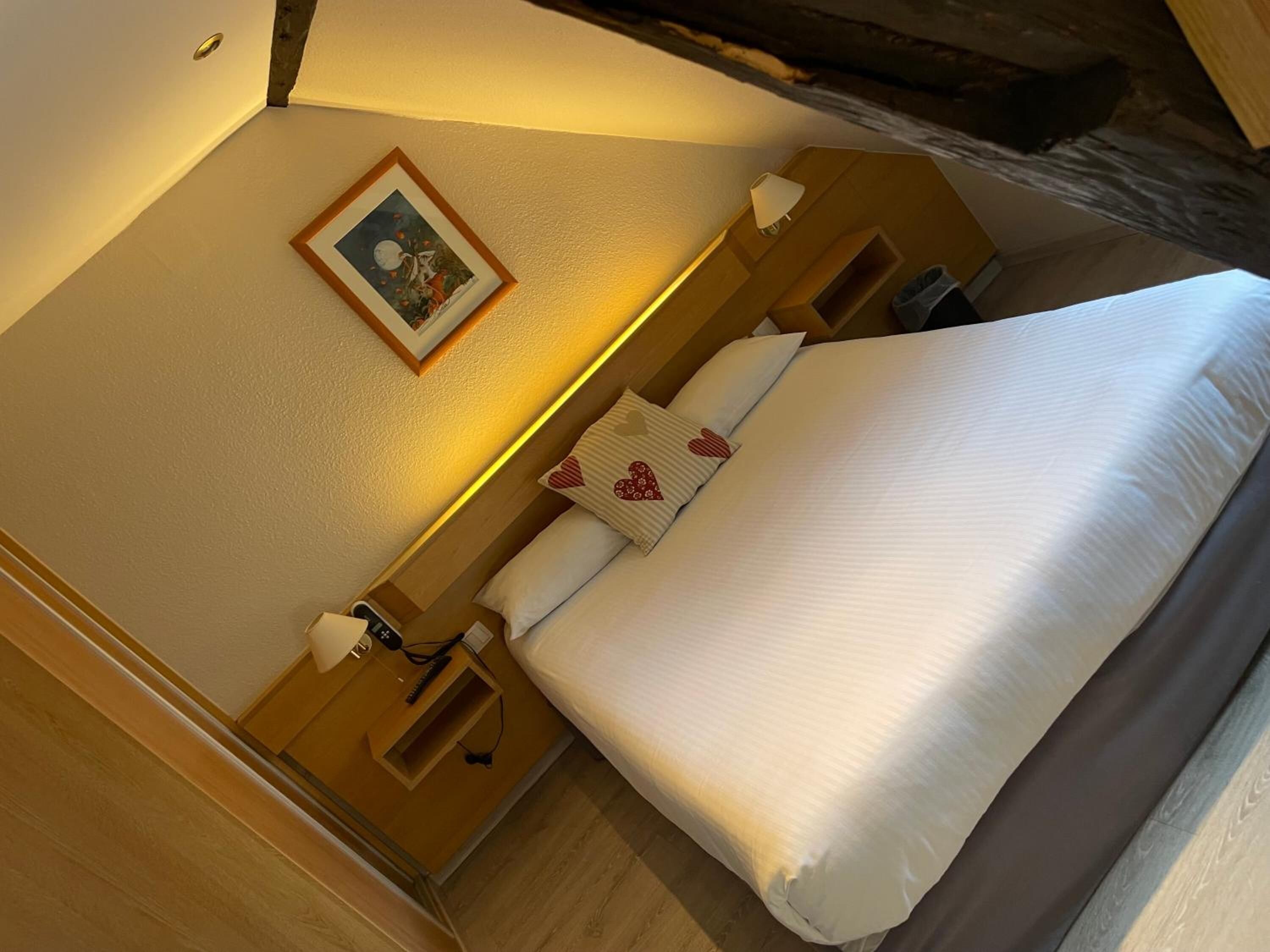 Una habitación de hotel con una cama bien hecha, una mesita de noche con lámpara, un enchufe y un pequeño cuadro en la pared.