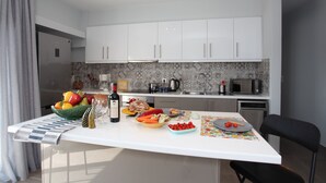A modern kitchen with white cabinets, a central island with a variety of fruits and vegetables, a wine bottle, and a microwave. - Edem apartment Sea View (Palaio Faliro)