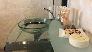 A bathroom with a glass bowl sink, a mirror, and toiletries including a body wash and a cream. - Tashatha Hotel (Tutume)