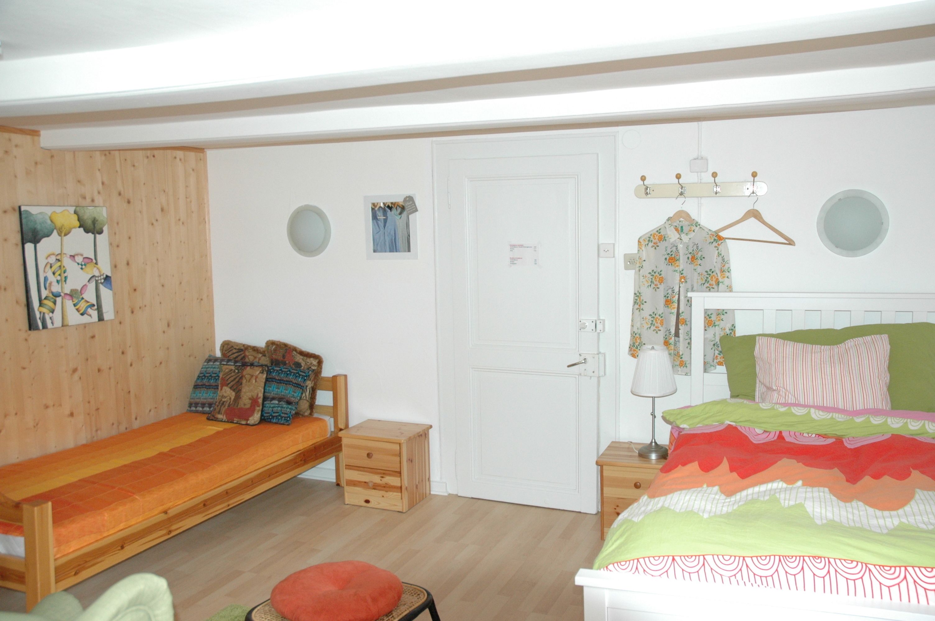 A bedroom with a wooden bed, a wooden bench, a small wooden chest, a red cushion, a white door, and a wall-mounted coat rack with a floral shirt.