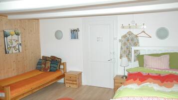 A bedroom with a wooden bed, a wooden bench, a small wooden chest, a red cushion, a white door, and a wall-mounted coat rack with a floral shirt.