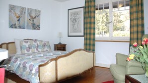 Un dormitorio con cama, mesita de noche, silla, ventana con cortinas y obras de arte en las paredes.