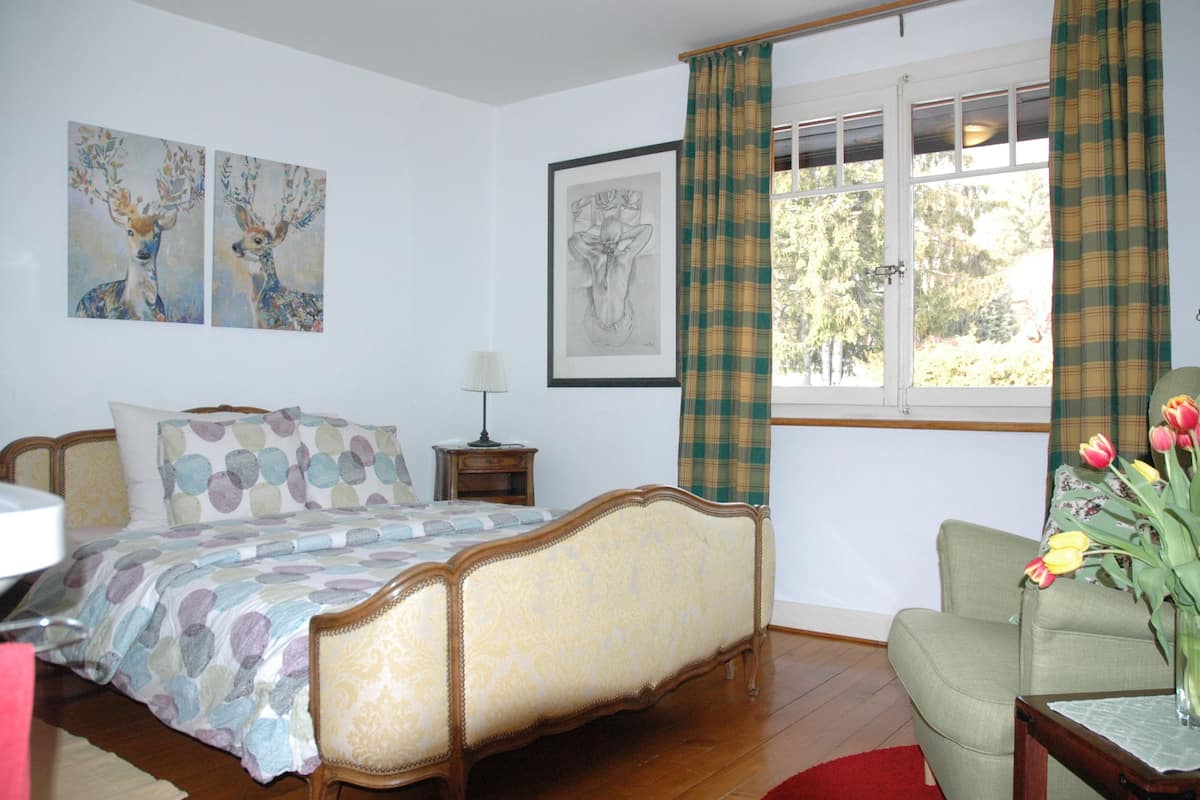 A bedroom with a bed, a nightstand, a chair, a window with curtains, and artwork on the walls.