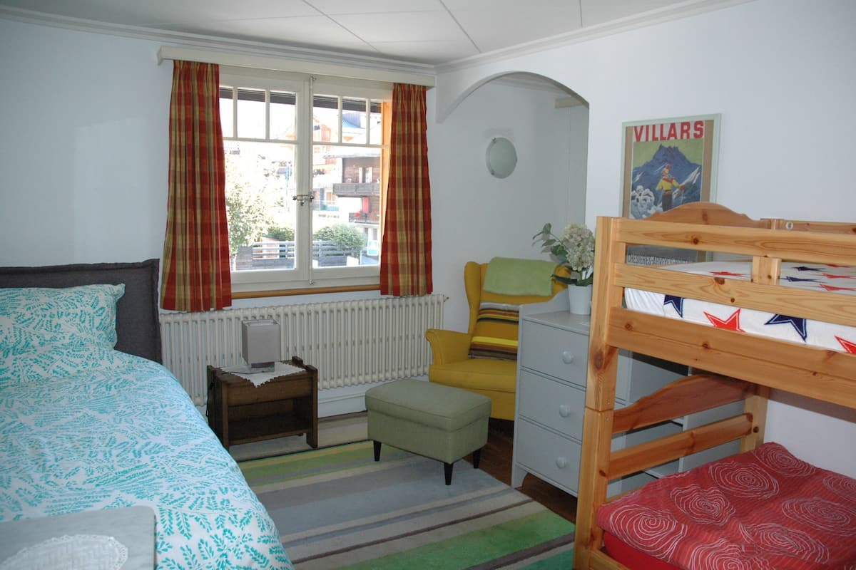 A bedroom with a bunk bed, a single bed, a radiator, a window with curtains, a small table with a lamp, and a poster on the wall.
