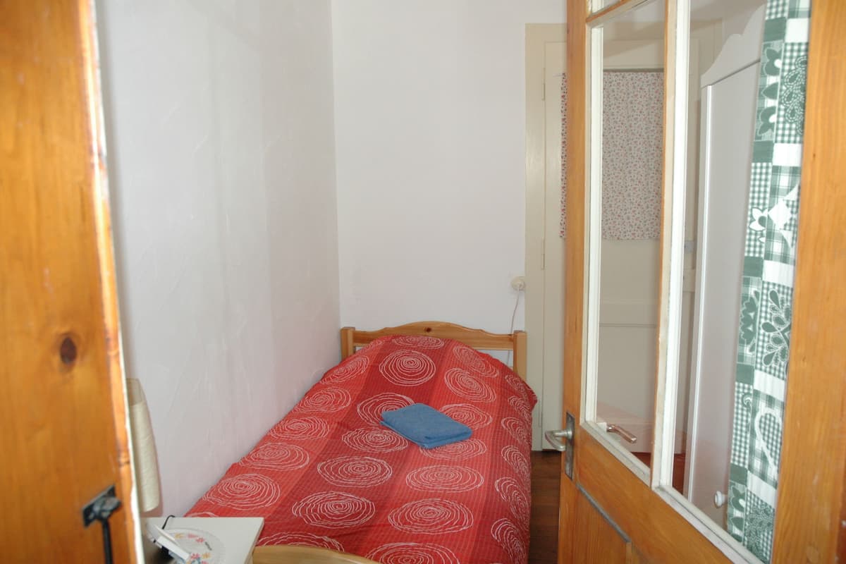 A small, narrow room with a single bed, a wooden door, and a window with a patterned curtain.