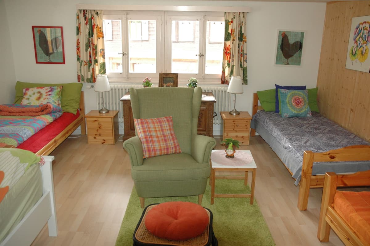 A cozy room with two beds, a green armchair, a small table, and a window with curtains.