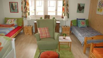 A cozy room with two beds, a green armchair, a small table, and a window with curtains.