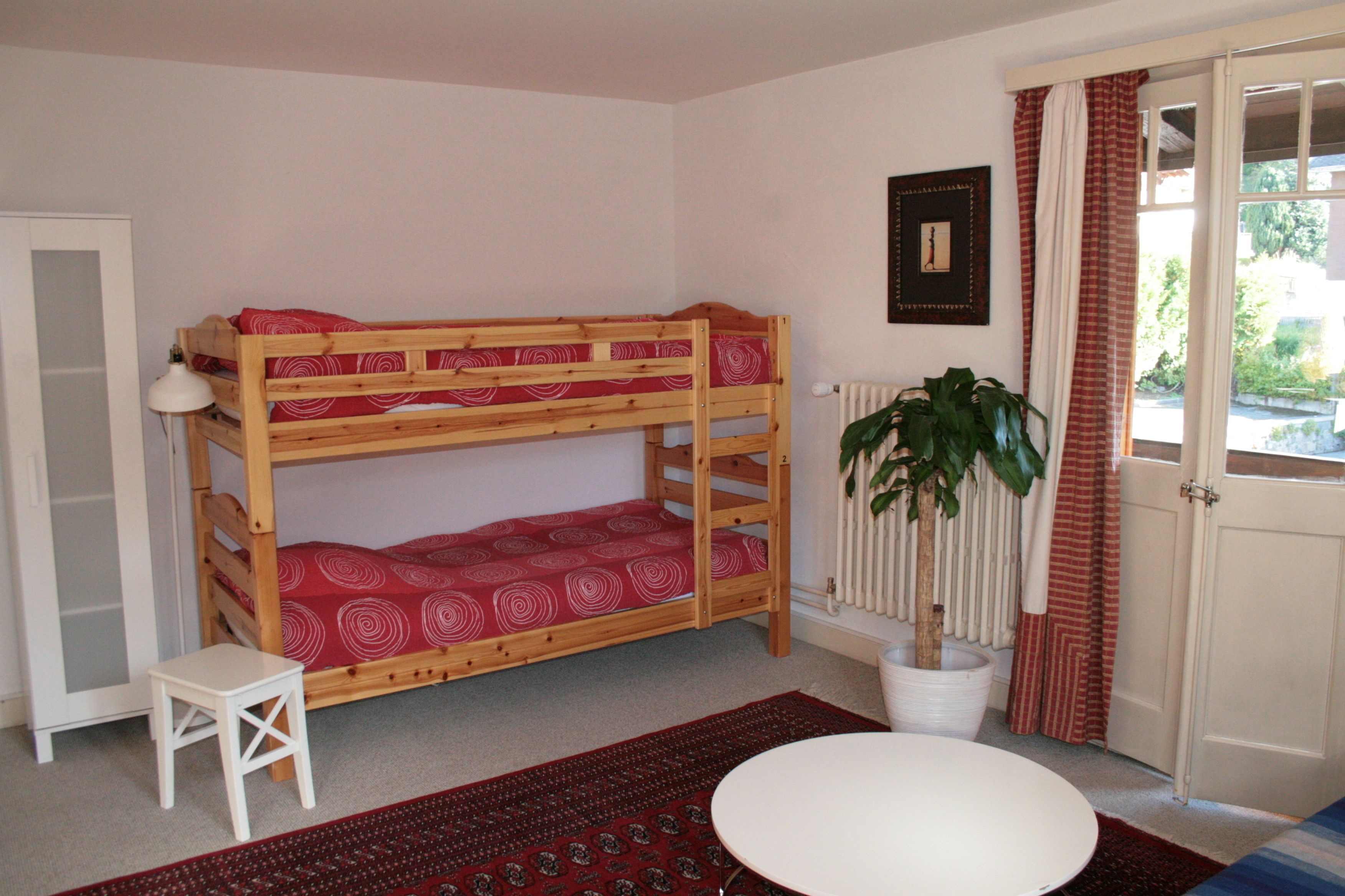 A bunk bed with a red patterned bedspread, a small white table, a potted plant, and a view through a door.