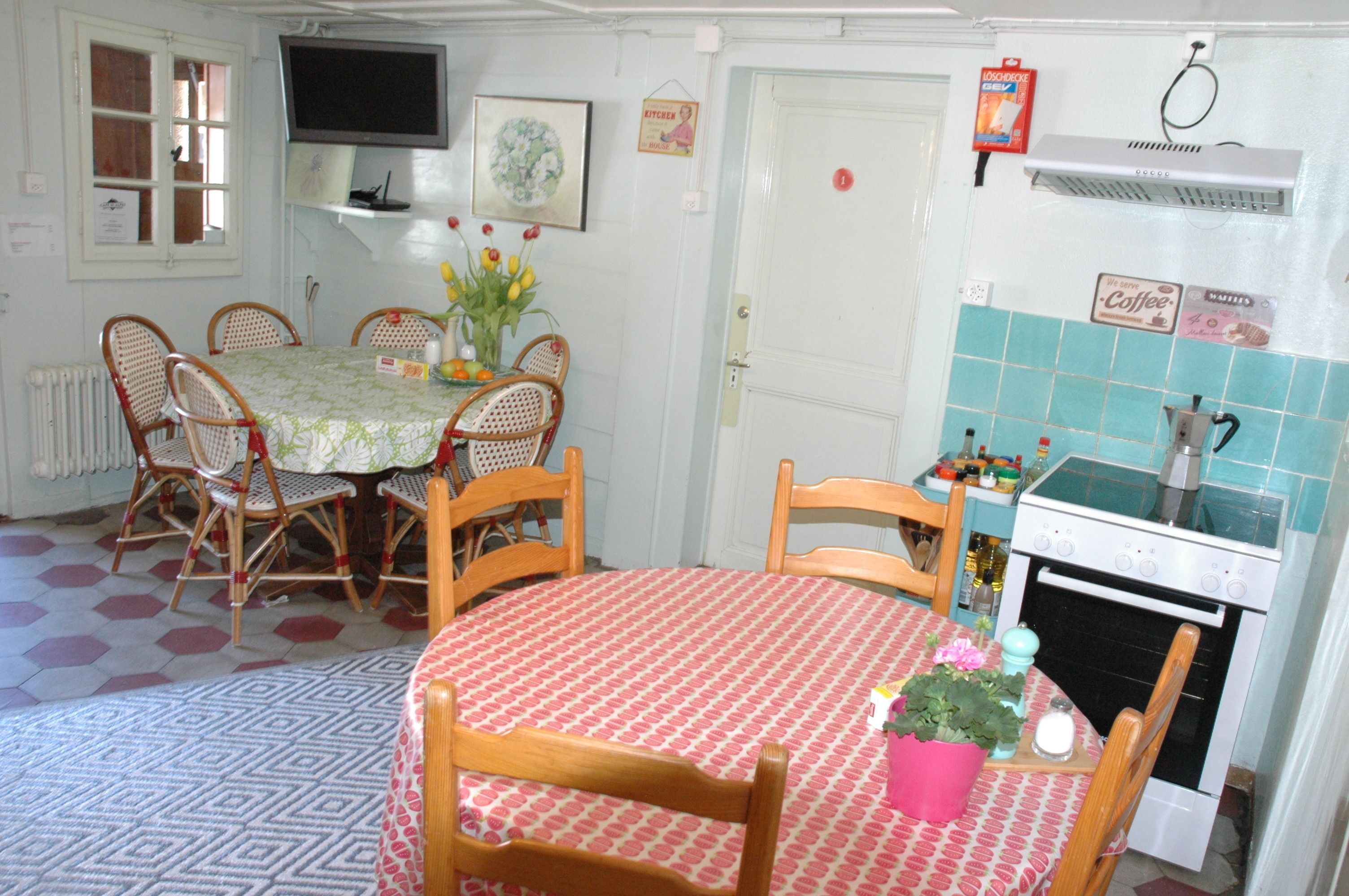 Un'area pranzo con un tavolo rotondo ricoperto da una tovaglia a quadretti rossi e bianchi, circondato da sedie in legno. Si nota un piccolo tavolo con tovaglia rosa e una pianta in vaso. Sullo sfondo, si intravede una zona cucina con un fornello e uno schienale piastrellato.