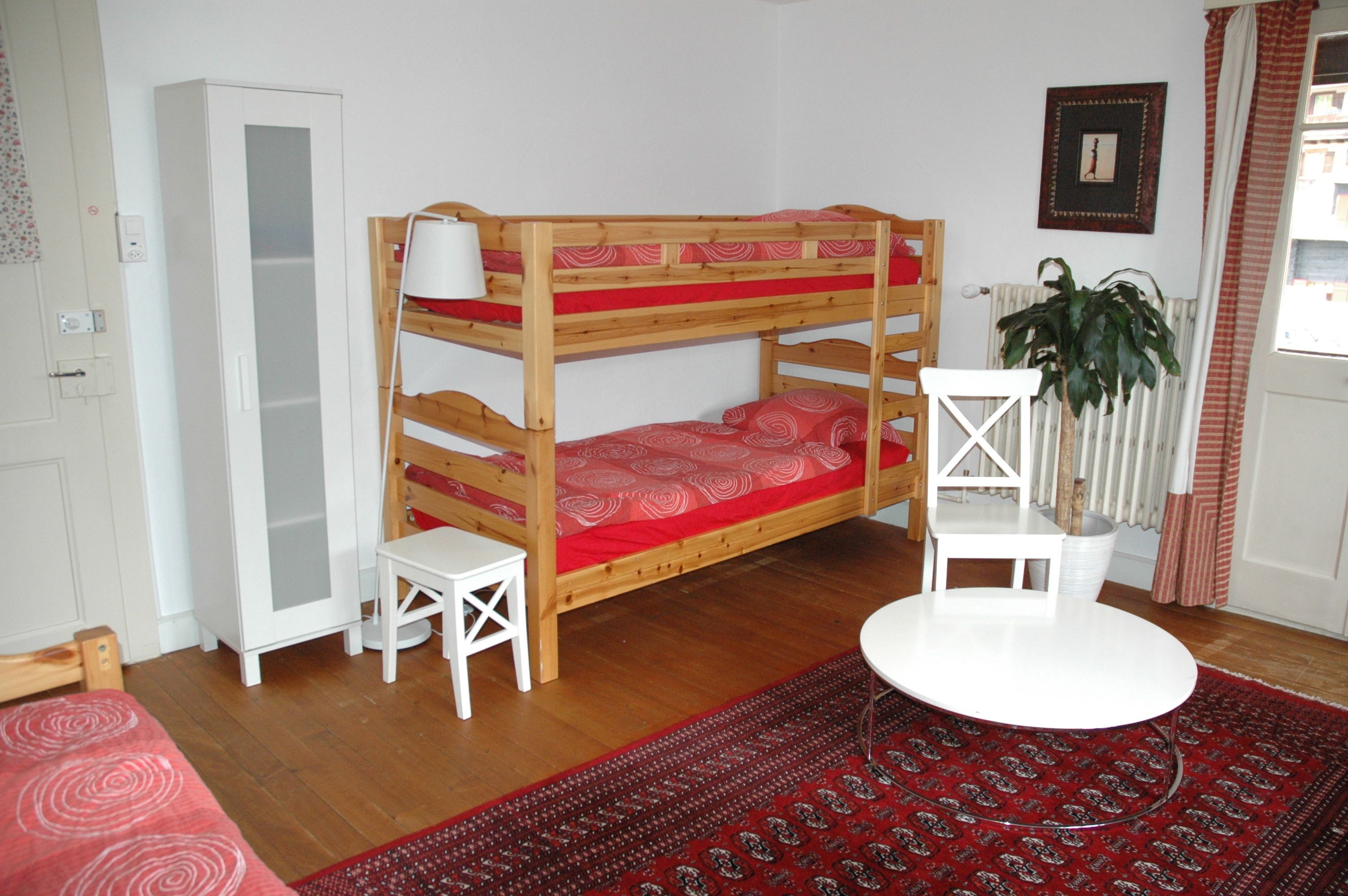 Un lit superposé avec un matelas rouge, un bureau blanc, une chaise blanche, un radiateur blanc et un tapis rouge à motifs.