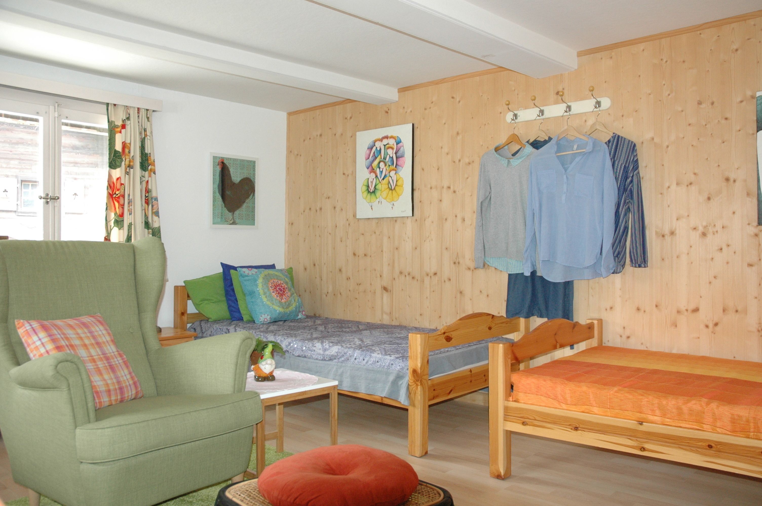 Een kamer met een groene fauteuil, een eenpersoonsbed, een klein tafeltje en een houten wand met een schilderij en kleding.