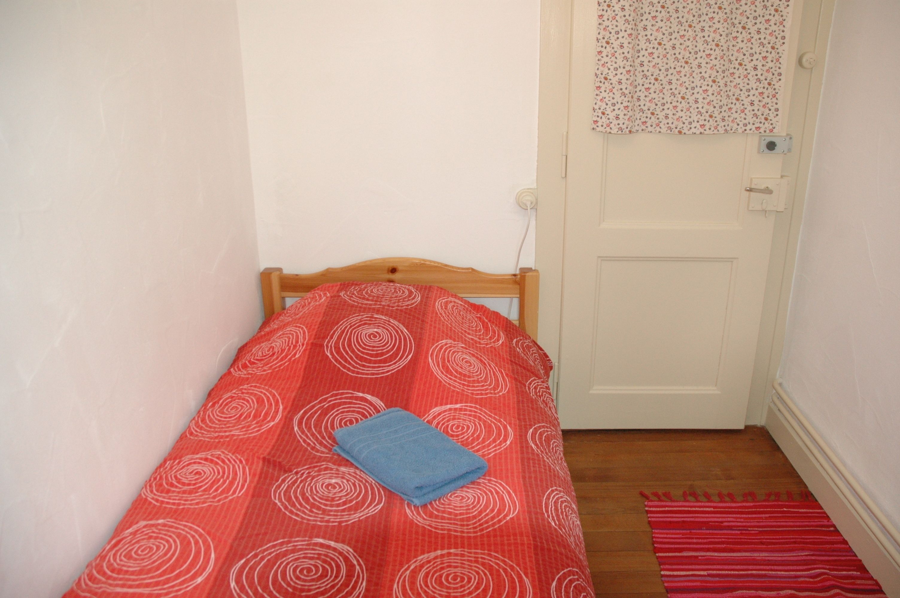 Una habitación pequeña con cama individual, cabecera de madera, colcha estampada en rojo y blanco, toalla azul y alfombra a rayas rojas sobre el piso.