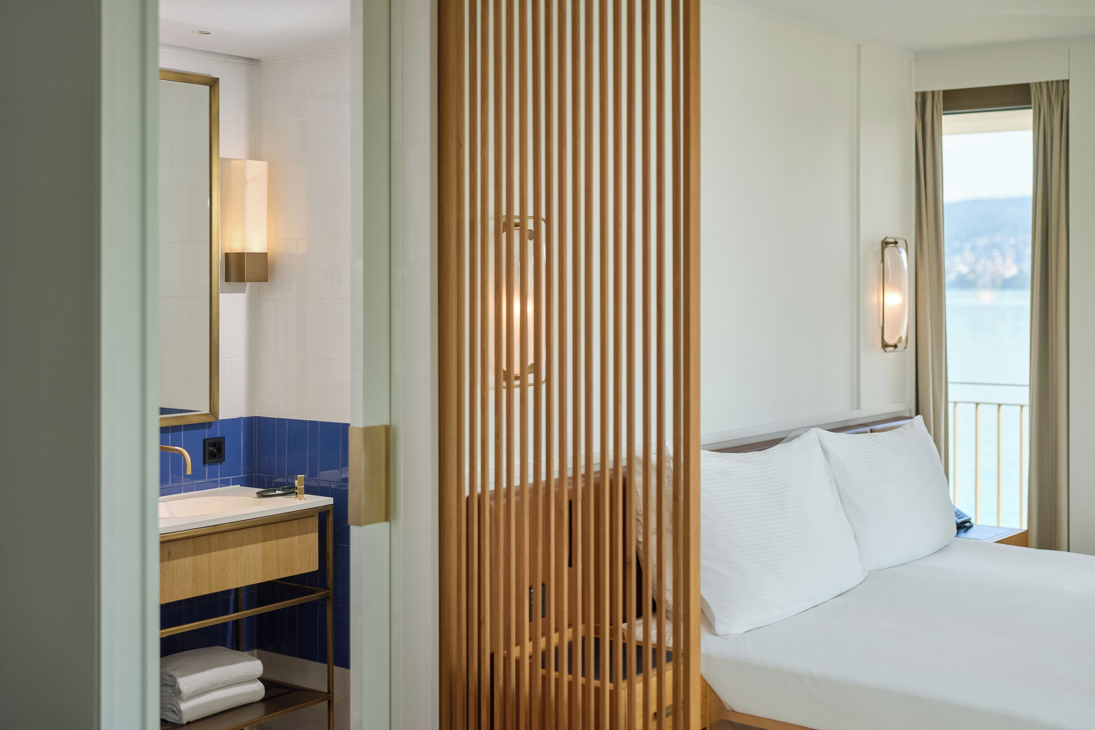 A hotel room with a wooden partition, a bed with white linens, a bathroom with a sink and mirror, and a view of the sea.