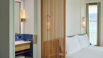A hotel room with a wooden partition, a bed with white linens, a bathroom with a sink and mirror, and a view of the sea.