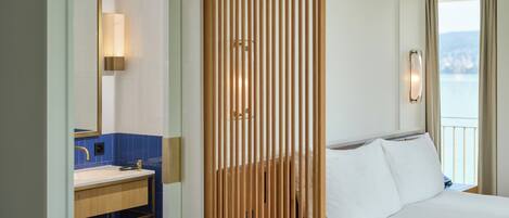 A hotel room with a wooden partition, a bed with white linens, a bathroom with a sink and mirror, and a view of the sea.