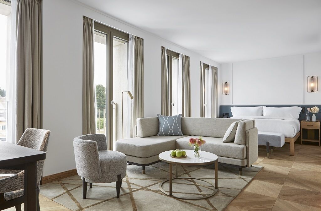 A modern hotel room with a sofa, armchair, round coffee table, and a bed with a headboard.